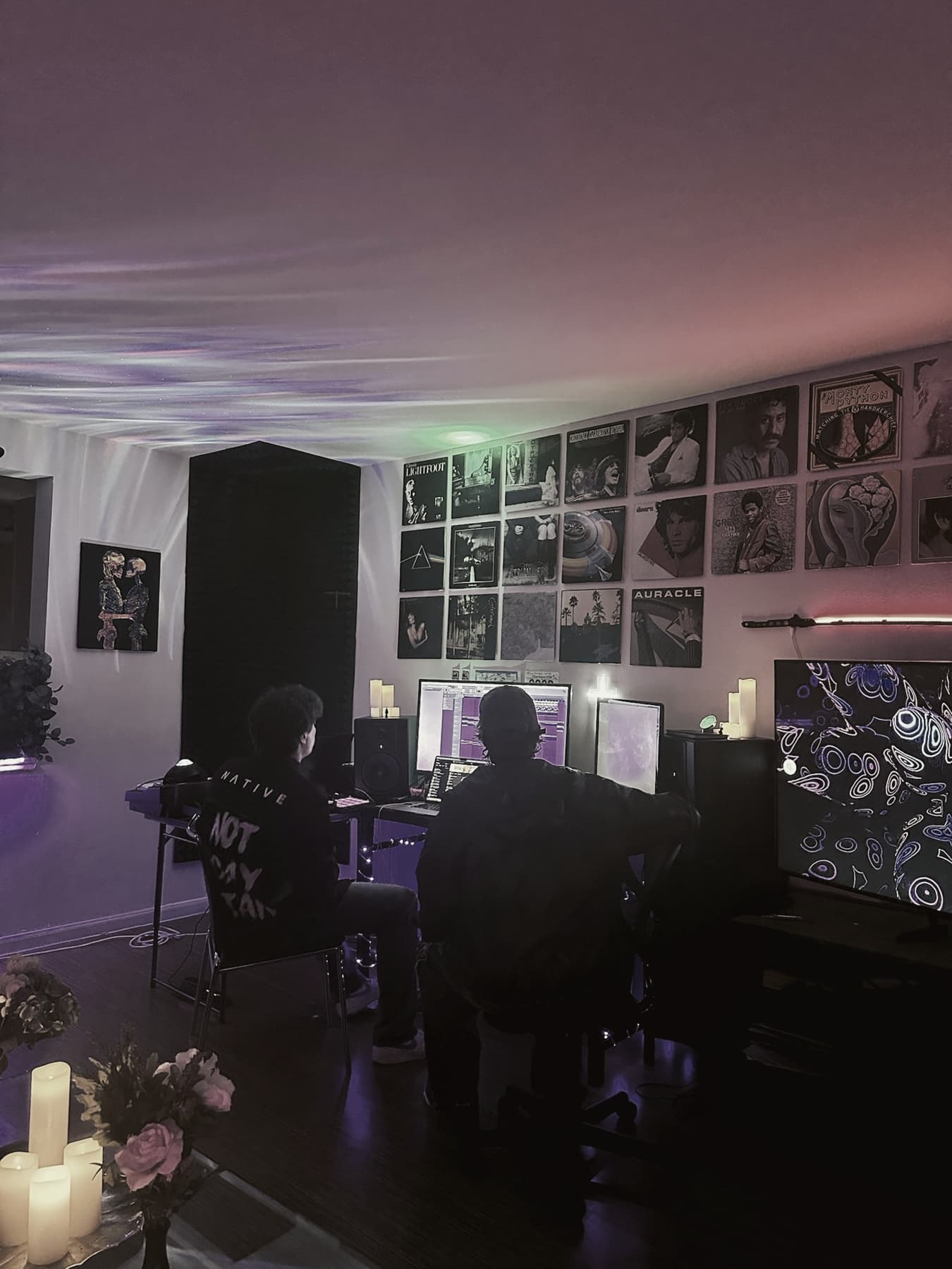 The studio from the back of the room: two people at the desk under the record wall, candles lit in the foreground
